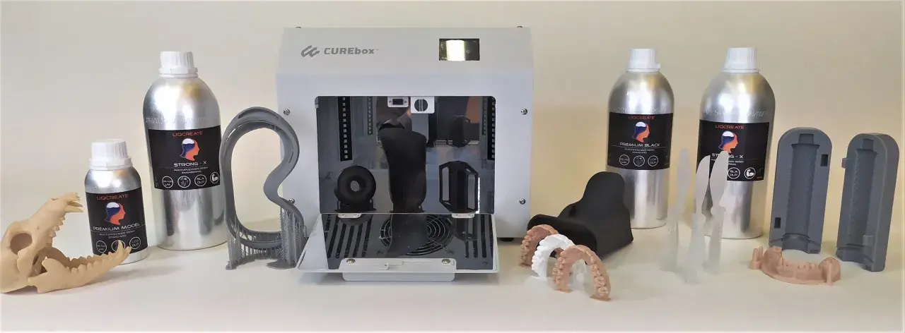 Flaconi di resina UV appiccicosa LIQCREATE, una stampante 3D CUREbox e vari modelli stampati, tra cui denti e un teschio.