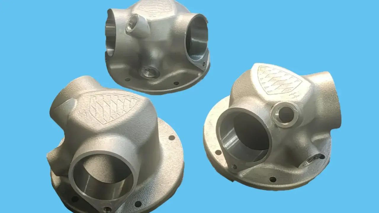 Tre componenti metallici complessi, realizzati con stampa 3D acciaio, mostrano un design intricato con diverse aperture e un logo inciso.