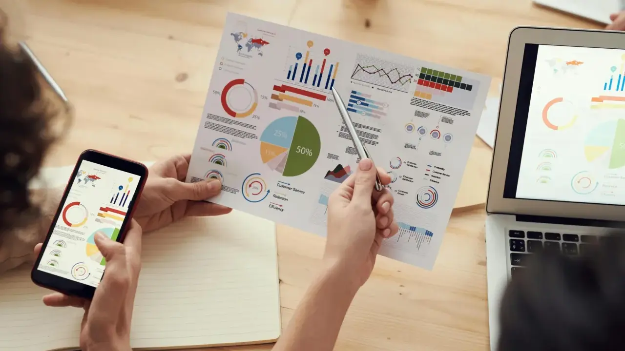 Persone analizzano grafici su fogli e smartphone, con un laptop aperto. Il software di programmazione aiuta a visualizzare dati complessi.