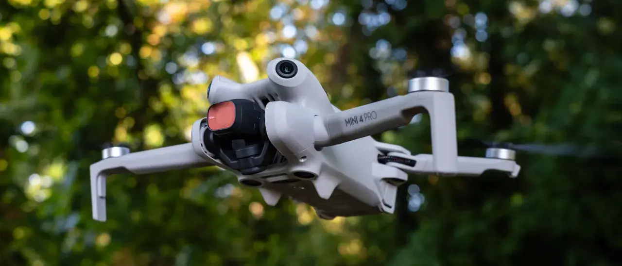Il DJI Mini 4 Pro in uscita, con filtro rosso sulla fotocamera, vola tra gli alberi.