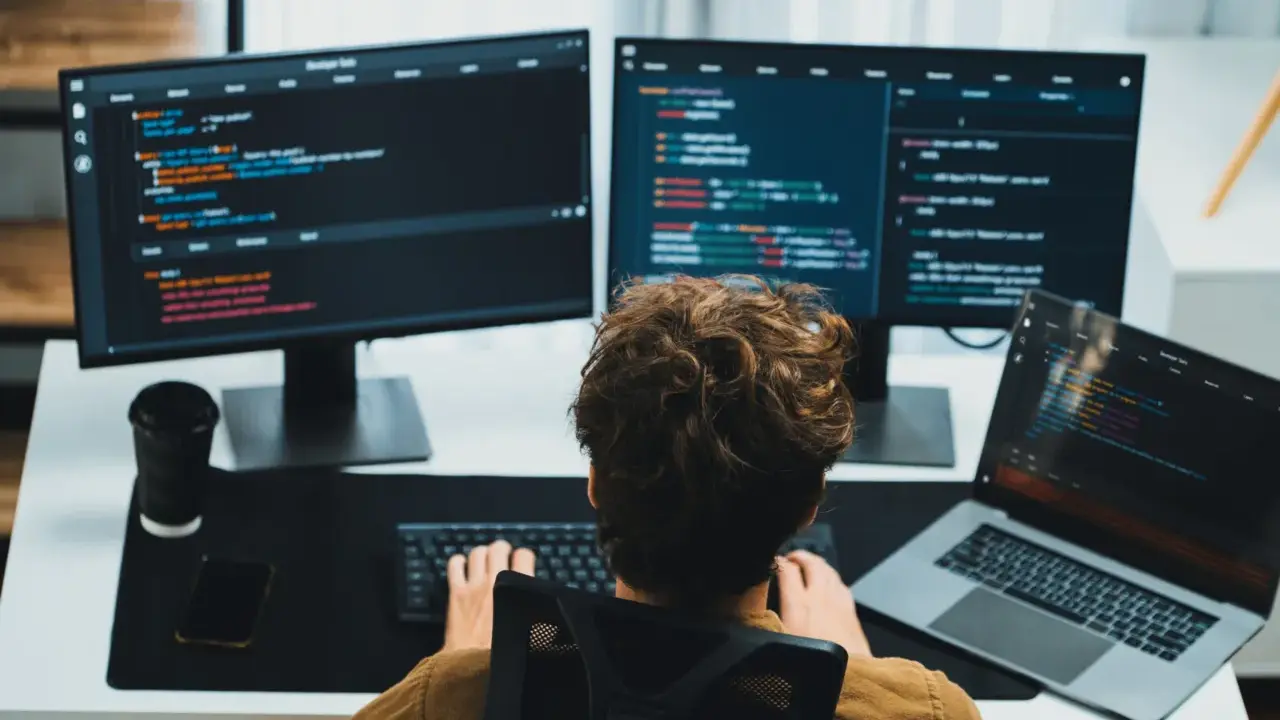 Un giovane con capelli ricci impara come diventare un programmatore, scrivendo codice su più monitor.