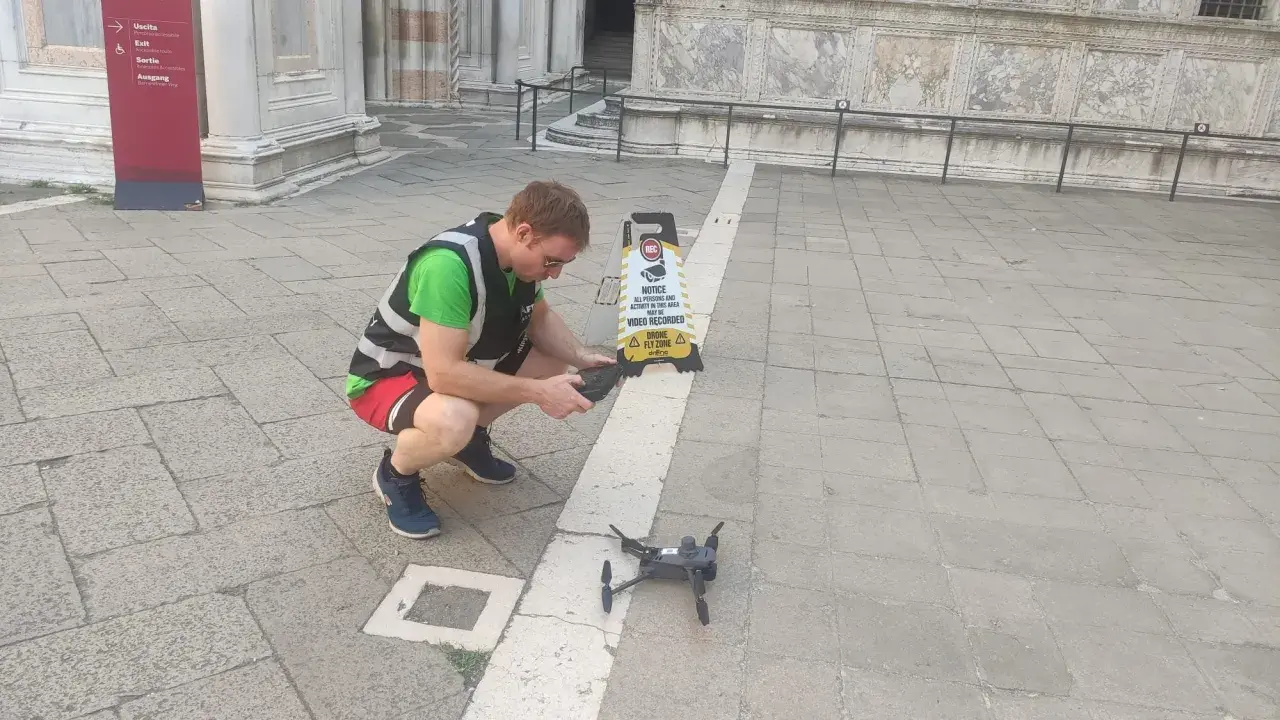 Uomo con giubbotto ad alta visibilità prepara un drone per riprese aeree, vicino a un cartello "Drone Fly Zone".