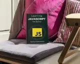 Libro "Learning JAVASCRIPT: The Basics" su un cuscino, con cuscini colorati e una sedia in legno sullo sfondo. Ideale per chi inizia la programmazione JavaScript.