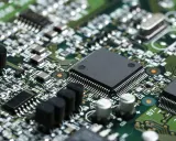 Primo piano di una scheda elettronica verde con numerosi componenti, tra cui un grande chip nero al centro, condensatori e resistori. Un'immagine dettagliata per il test componenti elettronici.