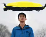 Un ragazzo guarda un drone che vola con la pioggia, tenendo un ombrello giallo sopra di lui.