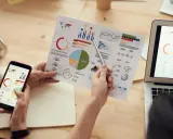 Persone analizzano grafici su fogli e smartphone, con un laptop aperto. Il software di programmazione aiuta a visualizzare dati complessi.