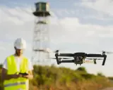 Uomo con elmetto e giubbotto alta visibilità controlla drone in volo. Potrebbe essere un corso per ottenere il patentino drone gratis Lussemburgo.