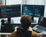Un giovane con capelli ricci impara come diventare un programmatore, scrivendo codice su più monitor.