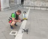 Uomo con giubbotto ad alta visibilità prepara un drone per riprese aeree, vicino a un cartello "Drone Fly Zone".