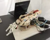 Mano robotica Arduino in legno e plastica, collegata a un laptop e alimentata da adattatori.
