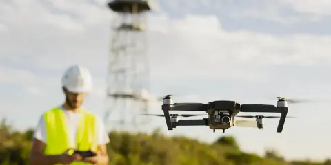 Uomo con elmetto e giubbotto alta visibilità controlla drone in volo. Potrebbe essere un corso per ottenere il patentino drone gratis Lussemburgo.