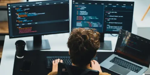 Un giovane con capelli ricci impara come diventare un programmatore, scrivendo codice su più monitor.