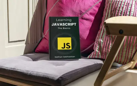 Libro "Learning JAVASCRIPT: The Basics" su un cuscino, con cuscini colorati e una sedia in legno sullo sfondo. Ideale per chi inizia la programmazione JavaScript.