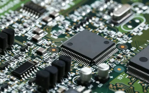 Primo piano di una scheda elettronica verde con numerosi componenti, tra cui un grande chip nero al centro, condensatori e resistori. Un'immagine dettagliata per il test componenti elettronici.