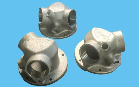 Tre componenti metallici complessi, realizzati con stampa 3D acciaio, mostrano un design intricato con diverse aperture e un logo inciso.