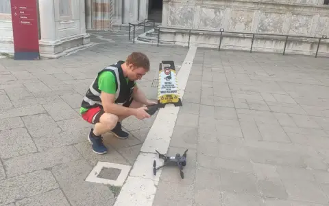 Uomo con giubbotto ad alta visibilità prepara un drone per riprese aeree, vicino a un cartello "Drone Fly Zone".