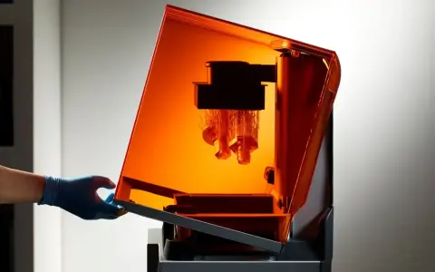 Mano con guanto blu solleva il coperchio arancione di una stampante 3D resina, rivelando oggetti in fase di stampa.