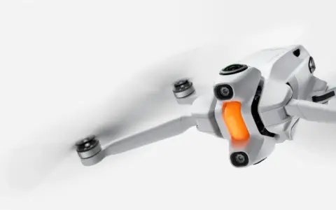 Un drone in volo, bianco e compatto, con eliche in rapido movimento e una luce arancione sul corpo.