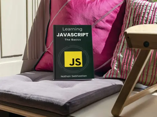 Libro "Learning JAVASCRIPT: The Basics" su un cuscino, con cuscini colorati e una sedia in legno sullo sfondo. Ideale per chi inizia la programmazione JavaScript.