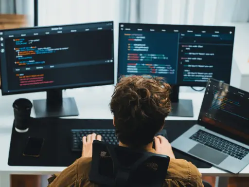 Un giovane con capelli ricci impara come diventare un programmatore, scrivendo codice su più monitor.