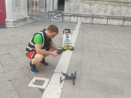 Uomo con giubbotto ad alta visibilità prepara un drone per riprese aeree, vicino a un cartello "Drone Fly Zone".