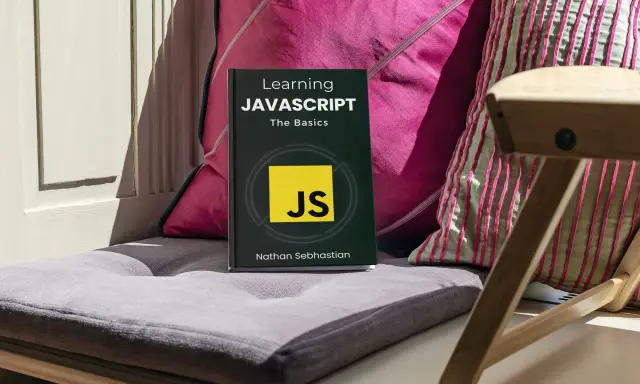 Libro "Learning JAVASCRIPT: The Basics" su un cuscino, con cuscini colorati e una sedia in legno sullo sfondo. Ideale per chi inizia la programmazione JavaScript.