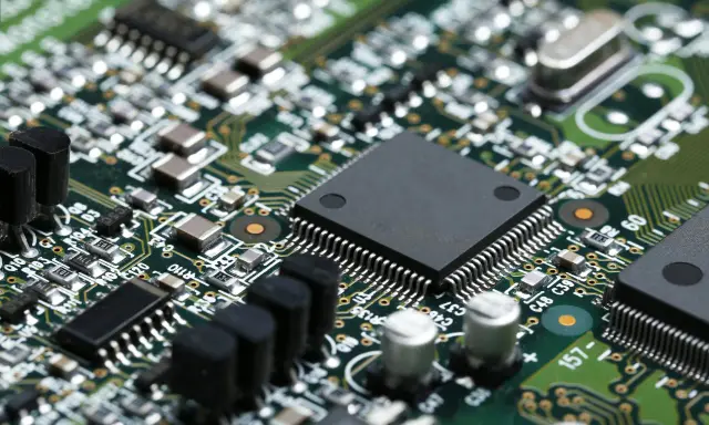 Primo piano di una scheda elettronica verde con numerosi componenti, tra cui un grande chip nero al centro, condensatori e resistori. Un'immagine dettagliata per il test componenti elettronici.