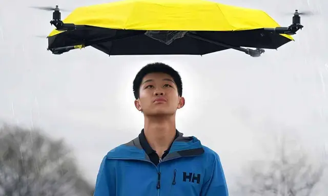 Un ragazzo guarda un drone che vola con la pioggia, tenendo un ombrello giallo sopra di lui.