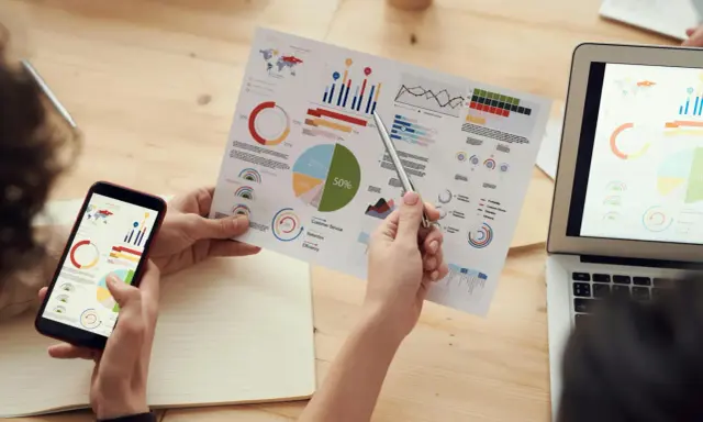 Persone analizzano grafici su fogli e smartphone, con un laptop aperto. Il software di programmazione aiuta a visualizzare dati complessi.
