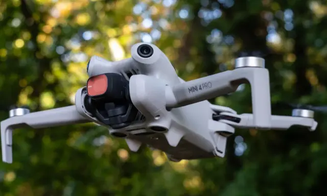 Il DJI Mini 4 Pro in uscita, con filtro rosso sulla fotocamera, vola tra gli alberi.
