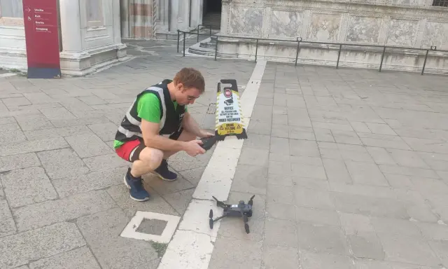 Uomo con giubbotto ad alta visibilità prepara un drone per riprese aeree, vicino a un cartello "Drone Fly Zone".