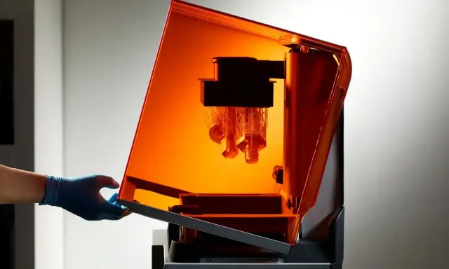 Mano con guanto blu solleva il coperchio arancione di una stampante 3D resina, rivelando oggetti in fase di stampa.