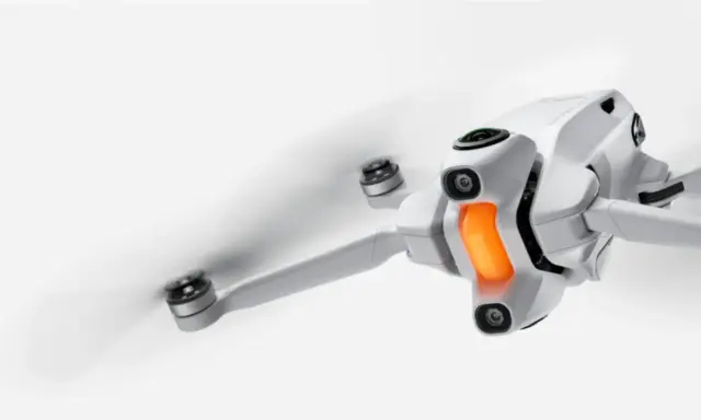 Un drone in volo, bianco e compatto, con eliche in rapido movimento e una luce arancione sul corpo.